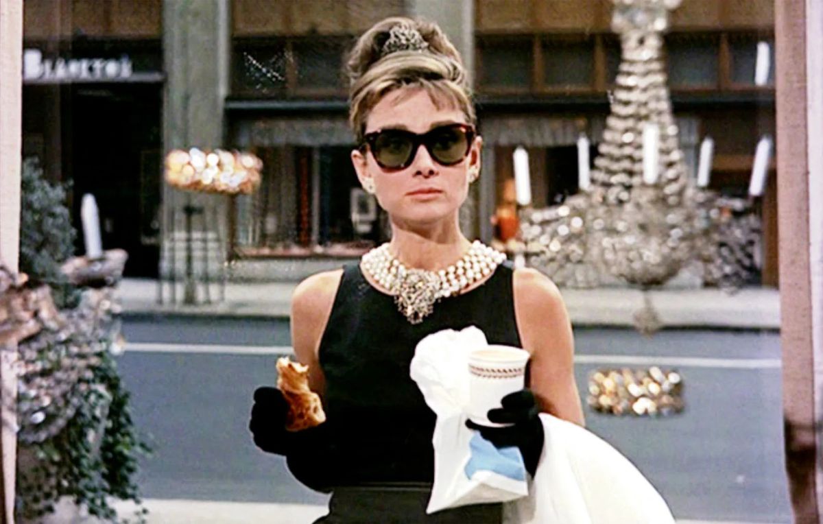 Holly Golightly, interprétée par Audrey Hepburn, dans Breakfast at Tiffany's, vêtue de sa robe noire emblématique, avec de grosses lunettes de soleil et un collier de perles. 