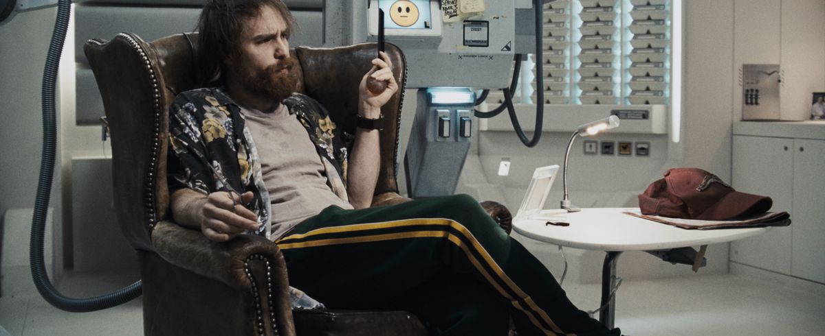 L'astronaute Sam Bell (Sam Rockwell), avec une grande barbe touffue, des cheveux hirsutes et des taches sur son t-shirt sale, affalé dans une chaise en cuir d'aspect antique au milieu des surfaces blanches et élégantes de la station spatiale et d'un robot carré arborant une non-expression smiley emoji dans la lune de Duncan Jones