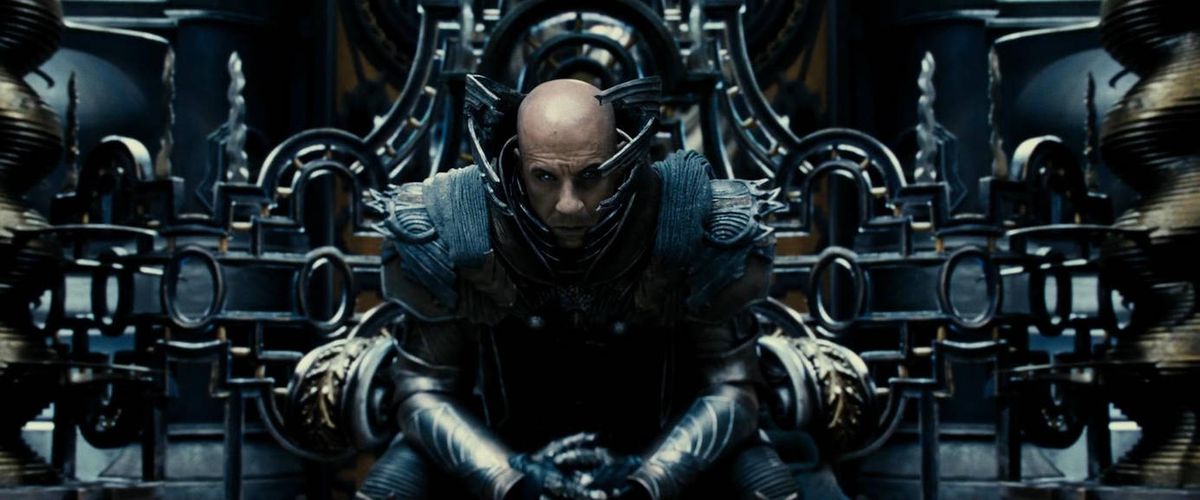 Un homme chauve (Vin Diesel) est assis sur un trône, vêtu d'une armure métallique élaborée et entouré d'une technologie élaborée sans but clair dans The Chronicles of Riddick