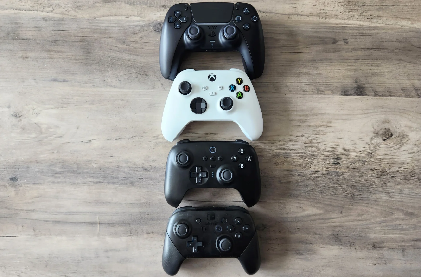 Quatre contrôleurs de jeux vidéo reposent sur une table en bois, l'un au-dessus de l'autre.  À partir du haut : la manette Sony DualSense PlayStation 5, la manette Microsoft Xbox Series X/S, la manette Bluetooth 8BitDo Ultimate et la manette Nintendo Switch Pro.