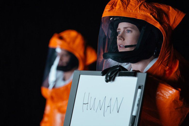 Amy Adams portant un costume de matières dangereuses orange et tenant un tableau effaçable à sec avec le mot «humain» écrit dessus dans Arrivée