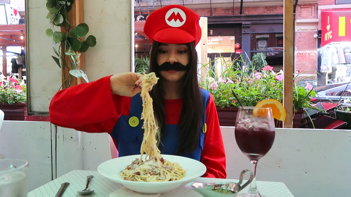Tina tient une énorme fourchette de spaghettis, les nouilles tombant de la fourchette.  Elle est habillée en Mario, assise dans une cabane à manger en plein air dans une rue ensoleillée et fleurie de New York.