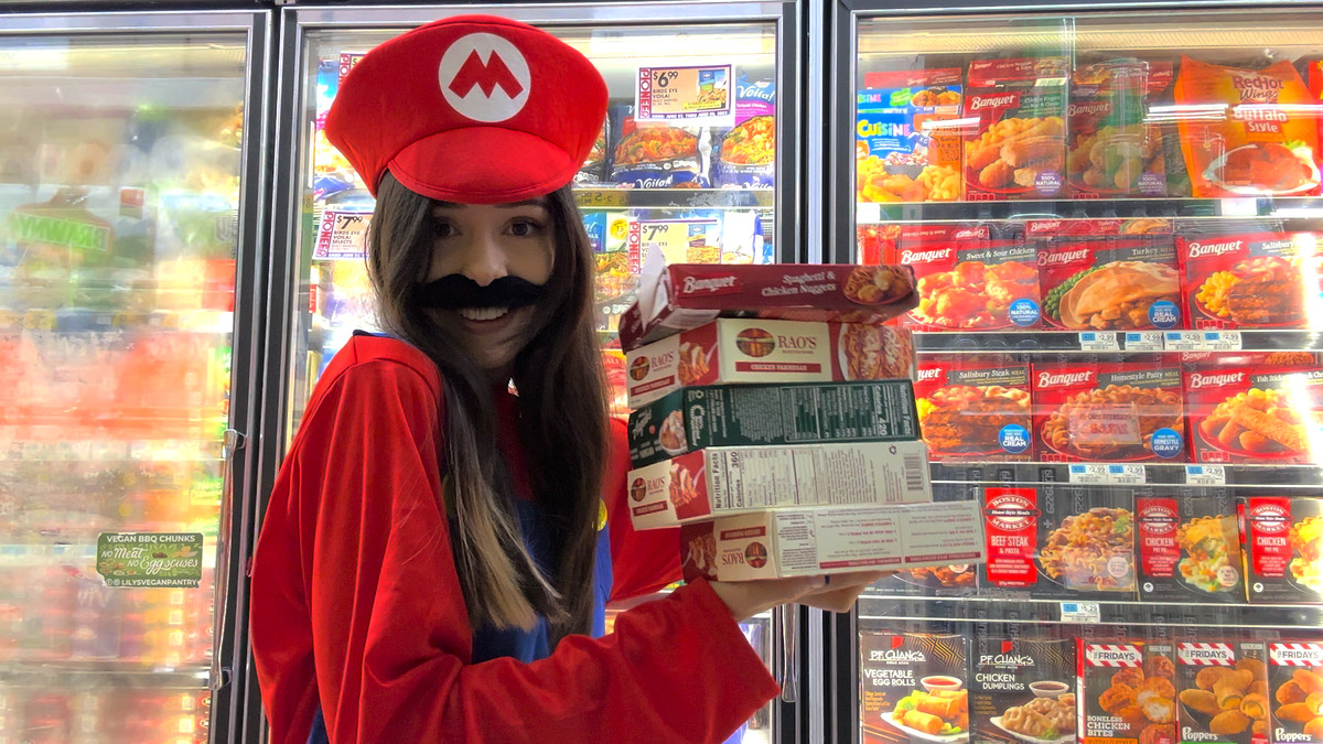 Tina, déguisée en Mario, tient joyeusement une pile de boîtes de spaghettis au micro-ondes, devant un grand congélateur d'aliments surgelés.