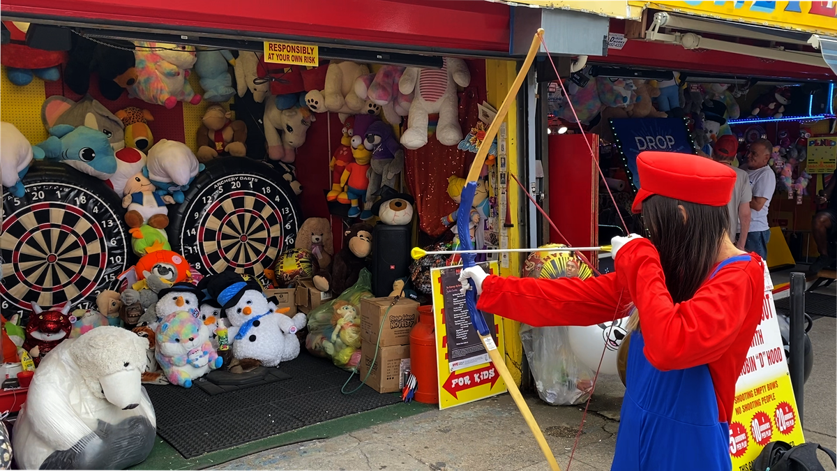 Tina, habillée en Mario, tenant mal un arc et une flèche en jouet, le pointant sur une cible dans un tas d'animaux en peluche.