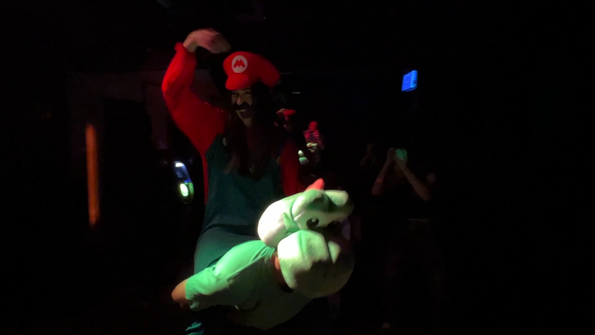 Tina déguisée en Mario, chevauchant un ami coiffé d'une tête de Yoshi, dans un club sombre.  Son poing est levé triomphalement.