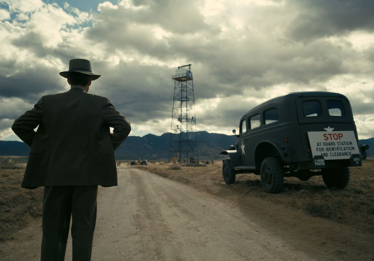 J. Robert Oppenheimer, en chapeau, les mains sur les hanches, se tient dos à la caméra et considère la tour sur le site d'essai de la bombe atomique devant un immense ciel nuageux du désert dans le film Oppenheimer