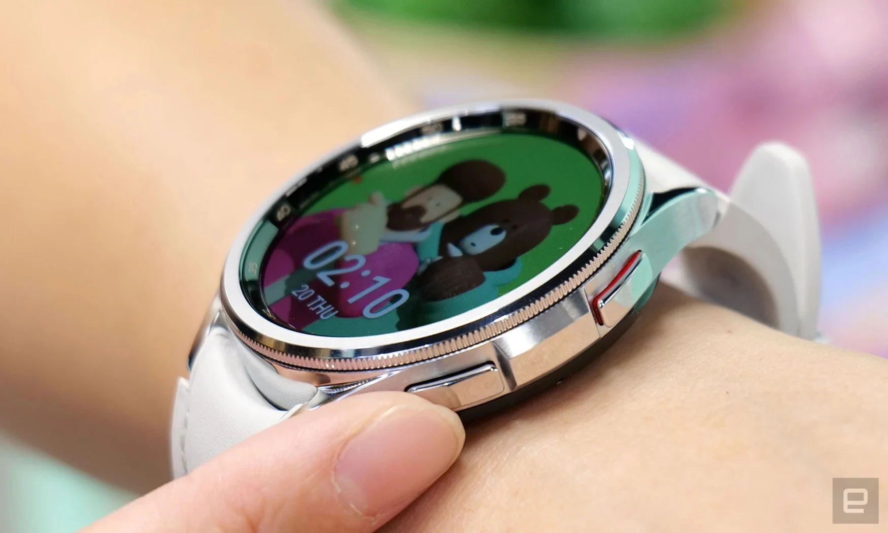 Photo en gros plan d'une montre intelligente Samsung Galaxy Watch 6 au poignet d'une personne.  Un doigt touche l'un des boutons matériels alors qu'une image d'un homme avec un ours en peluche s'affiche sur son écran.