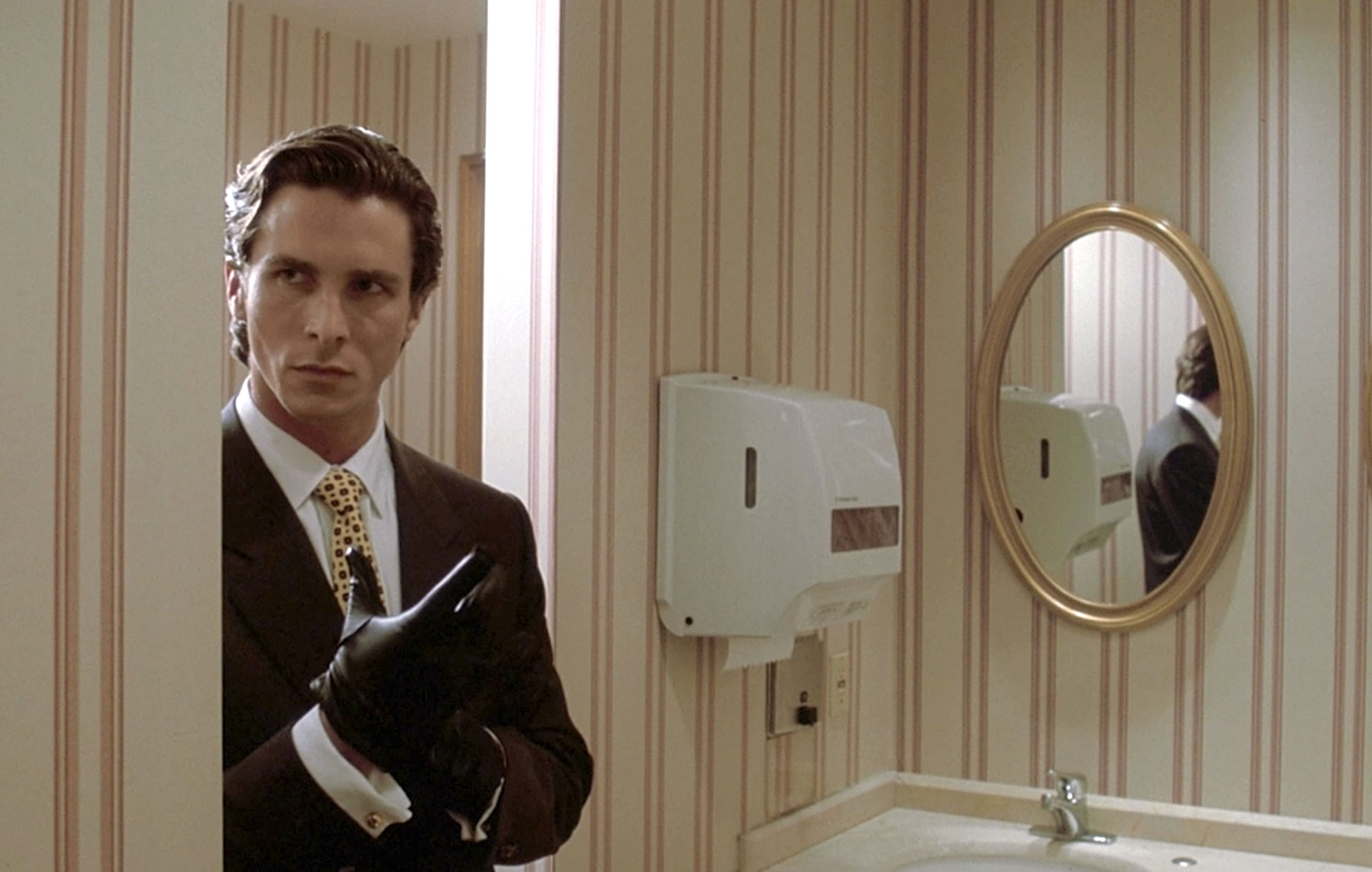 Patrick Bateman entre dans une salle de bain en enfilant un gant noir dans le film American Psycho.