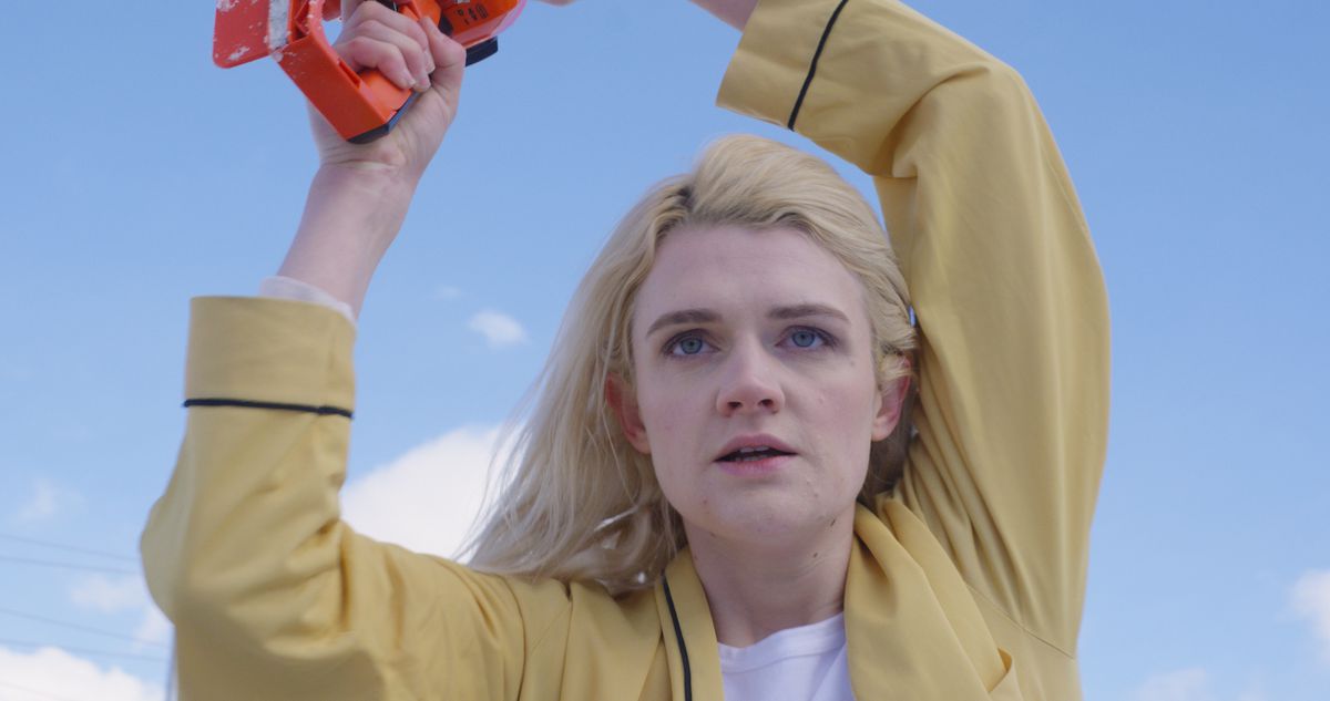 Gayle Rankin dans un manteau jaune tenant une tronçonneuse avec une poignée orange sur la tête dans Bad Things.