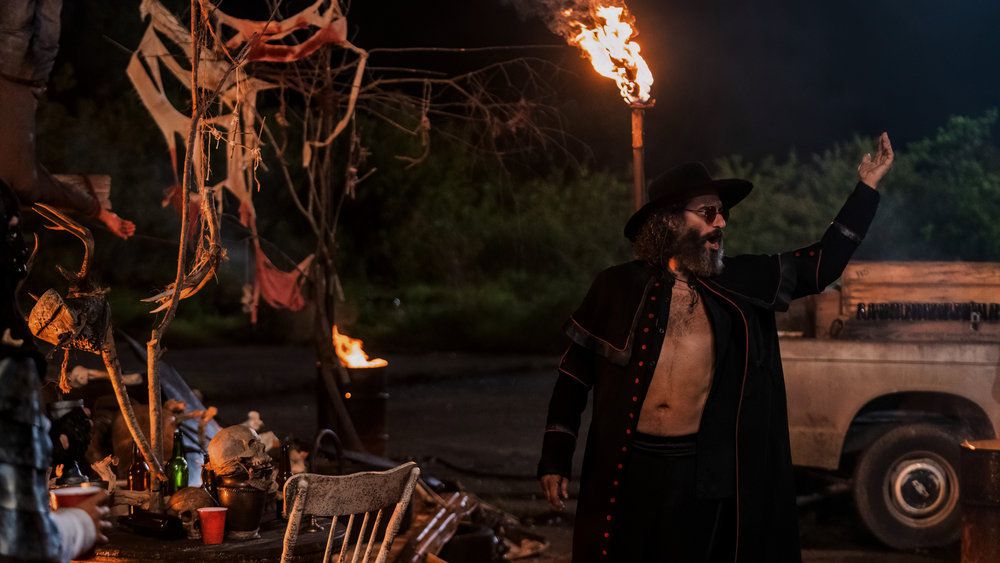 Jason Mantzoukas porte une veste déboutonnée, des lunettes de soleil et un chapeau noir avec son bras gauche levé en tant que chef des Holy Men, prédicateur dans Twisted Metal.  Il est entouré de ce qui ressemble à des détritus.