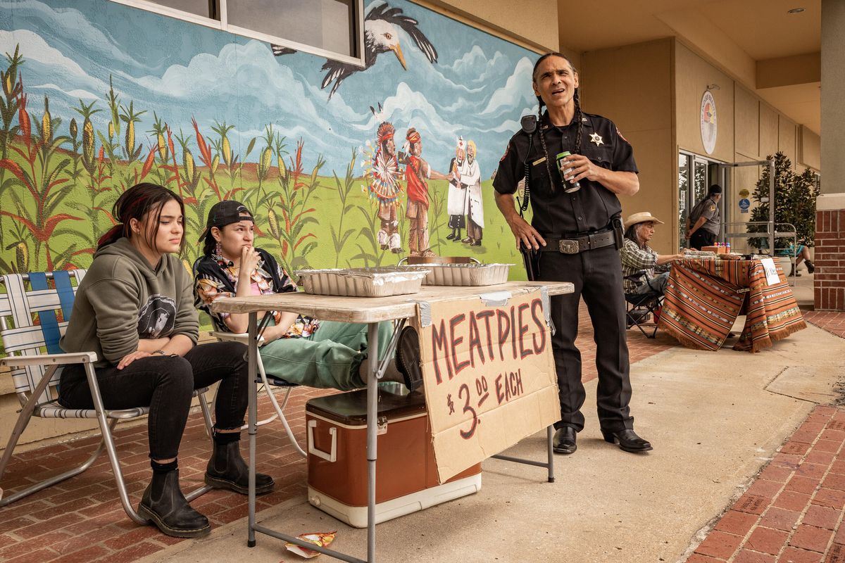 Devery Jacobs, Paulina Alexis et Zahn McClarnon se rassemblent autour d'une table annonçant des Meatpies à 3 $ chacun, devant une fresque murale, dans Reservation Dogs.