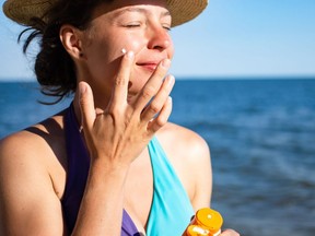 Lotion solaire SPF.  Belle femme souriante en chapeau d'été en paille appliquant de la crème solaire solaire à partir d'un récipient en plastique sur sa joue avec l'océan en arrière-plan, portant un maillot de bain bleu.