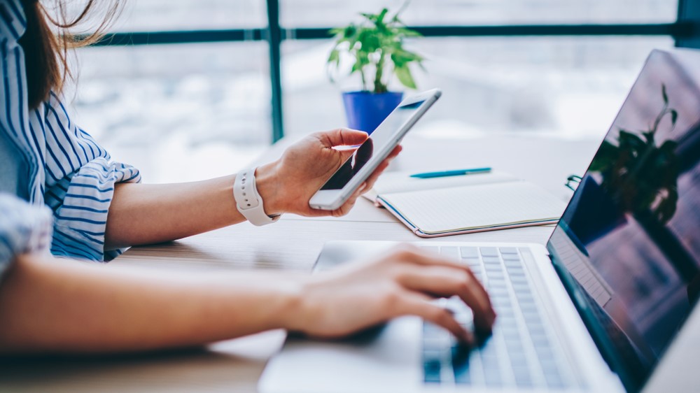 Une femme regardant un smartphone tout en utilisant un ordinateur portable