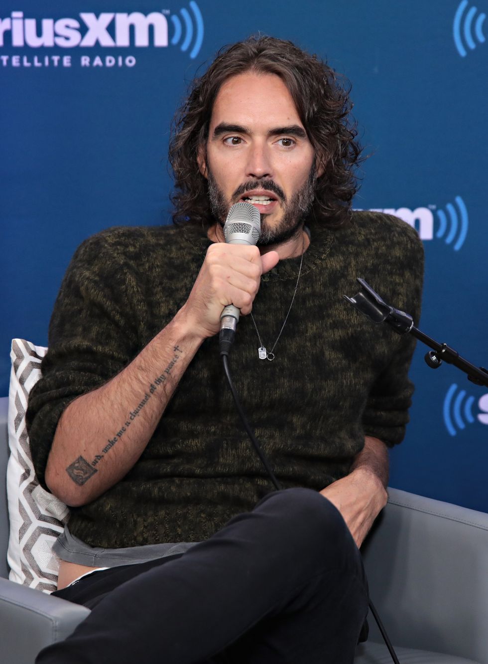 Russell Brand lors d'un événement en 2017, il est assis devant un fond bleu, parlant dans un microphone et portant un pull sombre