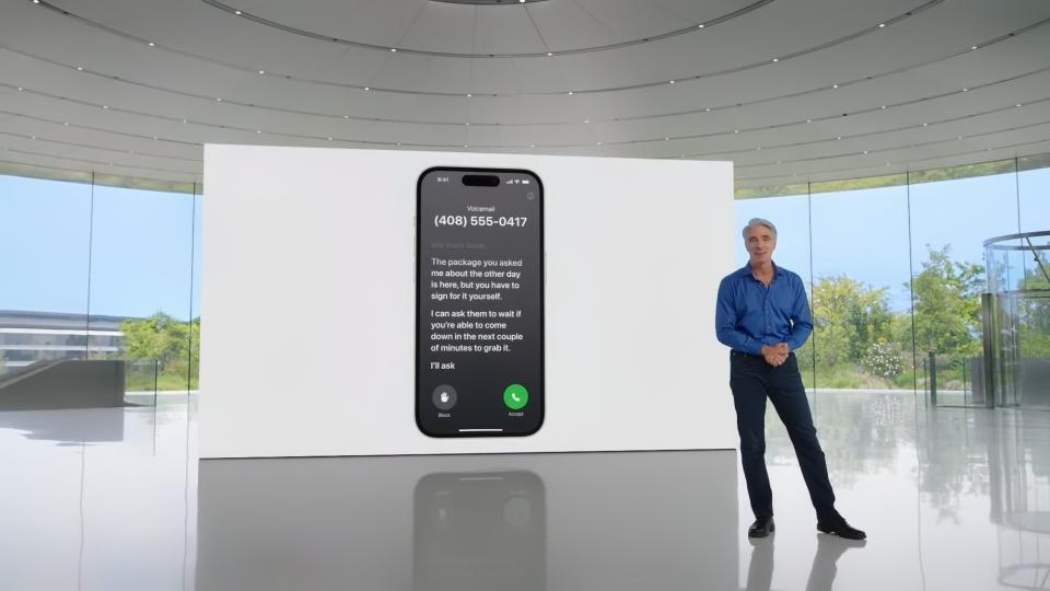 Craig Federighi, cadre d'Apple (chemise boutonnée bleue, pantalon noir) debout dans une pièce d'Apple Park (esthétique grise moderne avec des arbres visibles à l'extérieur des fenêtres).  Sur un écran derrière lui se trouve une démonstration de messagerie vocale en direct.