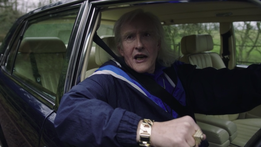 Jimmy Savile (Steve Coogan) vêtu d'un survêtement bleu de marque et penché par la fenêtre de sa voiture Bentley.