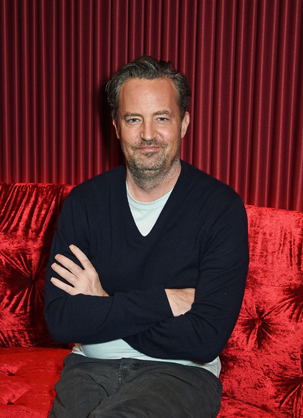 Matthew Perry est assis, les bras croisés, contre un canapé rouge avec un rideau rouge assorti derrière lui