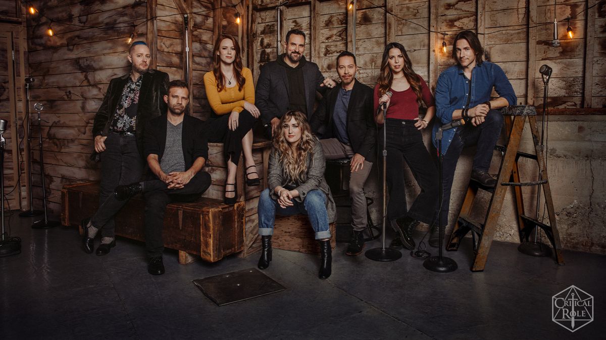 The cast of Critical Role in a rustic wooden building, standing around or sitting on crates with microphones surrounding them