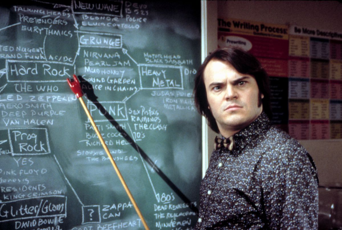 Jack Black donne à la caméra un regard super sournois alors qu'il se tient devant un tableau et pointe un pointeur vers une répartition des styles et sous-genres de la musique rock dans le film School of Rock de Richard Linklater en 2003.