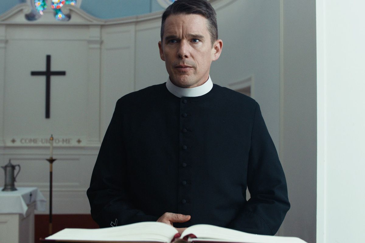 Un homme en tenue de prêtre (Ethan Hawke) se tient derrière un pupitre avec une croix noire visible en arrière-plan.