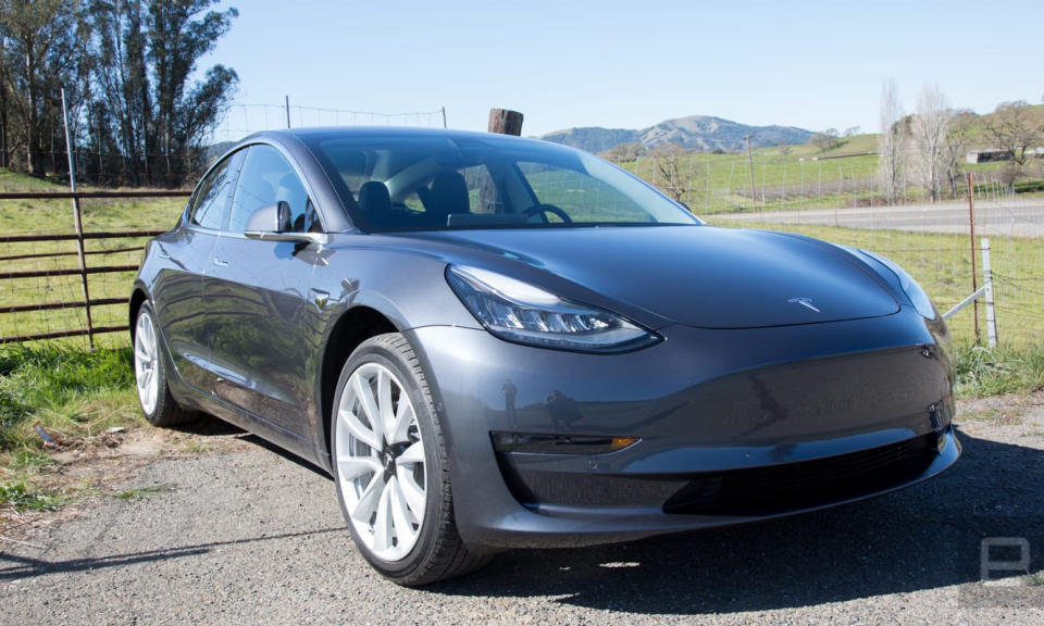 Photo de la Tesla Model 3 assise à l’extérieur, à côté d’un champ.  L'herbe verte, les arbres et les collines sont visibles au loin.