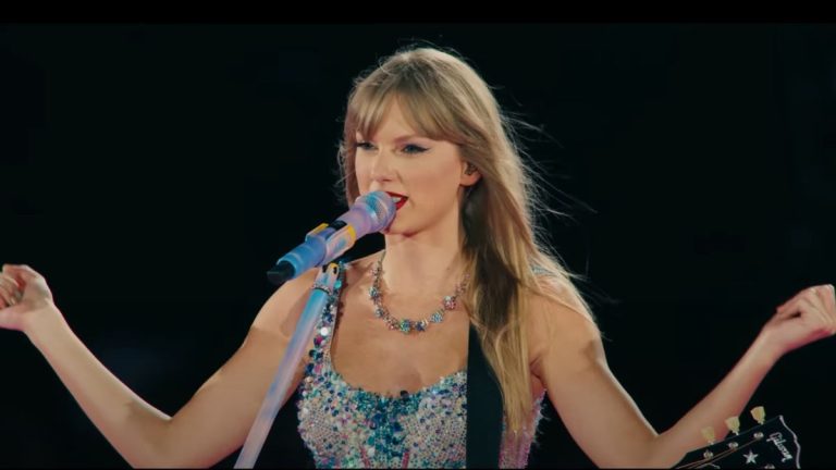 A screenshot from the Eras Tour trailer of Taylor Swift talking into a microphone and holding her arms up.