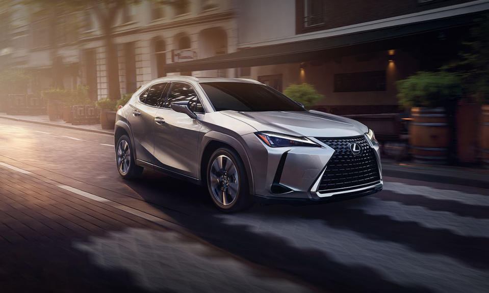 Photo marketing du véhicule hybride Lexus UX.  Le modèle argenté circule dans une rue de la ville avec un éclairage spectaculaire.