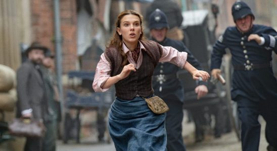 A press image of Millie Bobby Brown as Enola being chased by cops down a street in Enola Holmes 2.