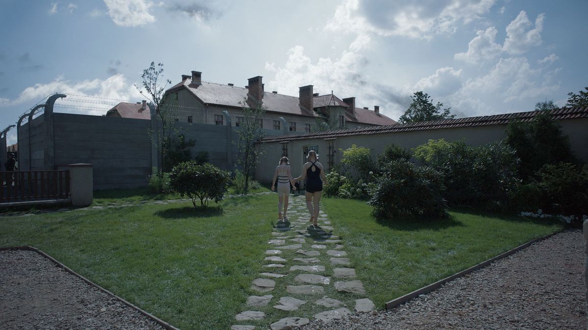 Deux jeunes filles marchent sur un chemin dans un jardin clos avec les pensions d'Auschwitz au-dessus d'elles dans la zone d'intérêt