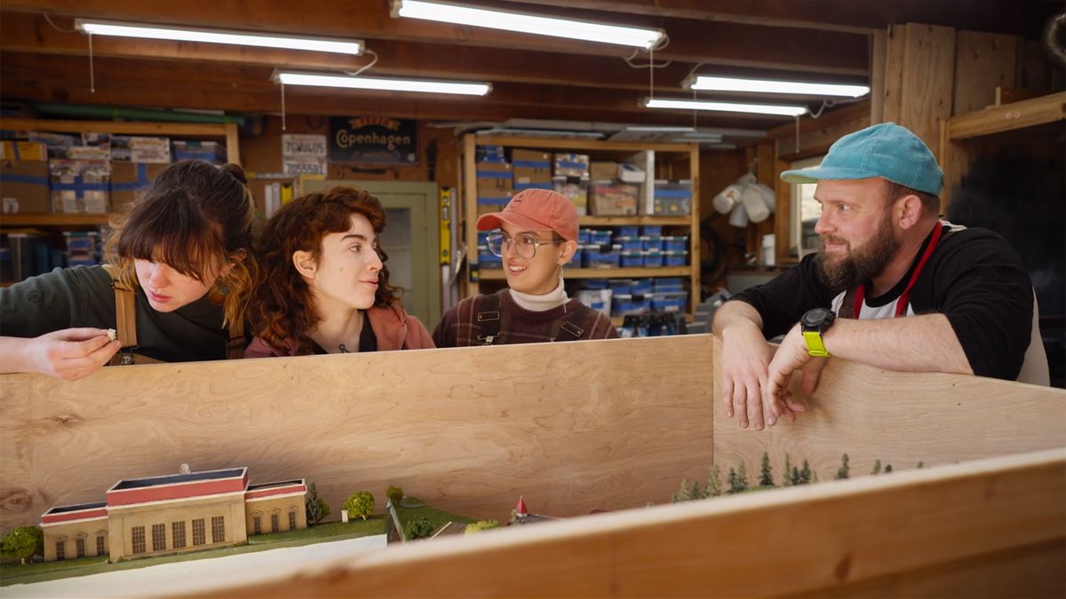 Rick Perry, portant une casquette bleue, se tient à côté de trois de ses coéquipiers dans un atelier grossièrement taillé avec des poutres apparentes.  Des bacs de miniatures sont posés sur des étagères en arrière-plan.