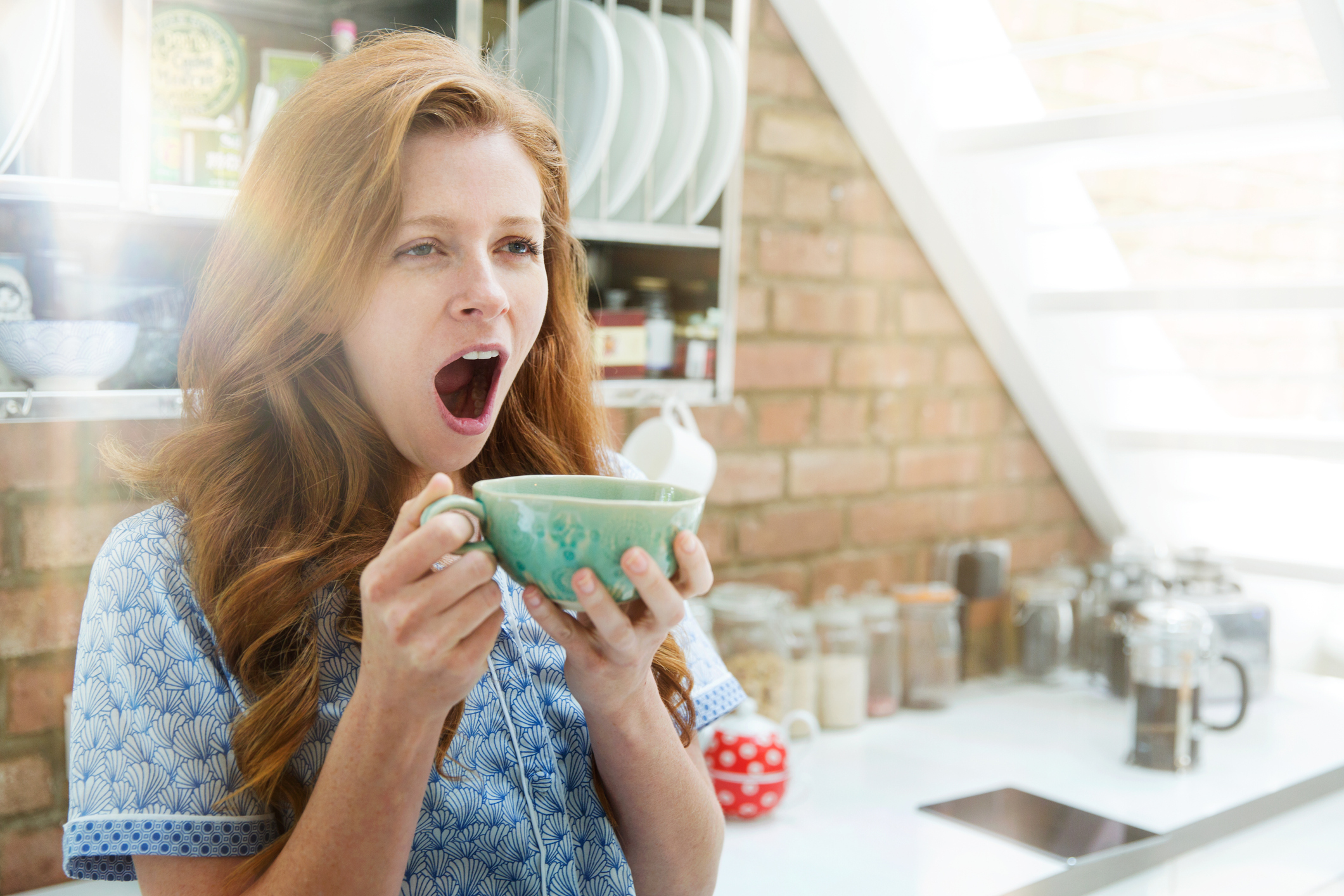 Une femme aux cheveux rouges bâille tout en tenant une tasse à deux mains