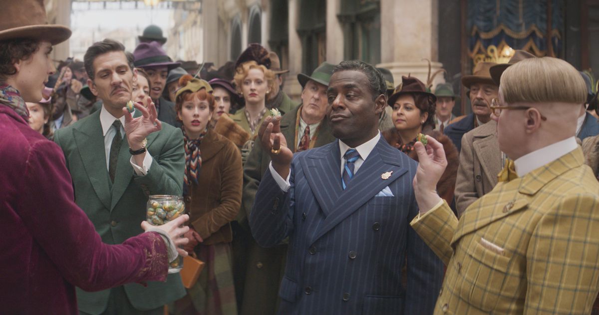 Mathew Baynton en costume vert, Paterson Joseph en costume bleu et Matt Lucas en costume jaune parlant à Willy Wonka