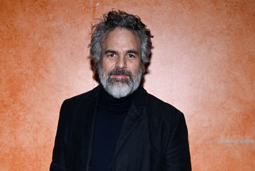 Mark Ruffalo, un homme aux cheveux gris et noirs et à la barbe grise et noire, portant un pantalon noir, un manteau noir et un col roulé noir, pose devant un mur orange