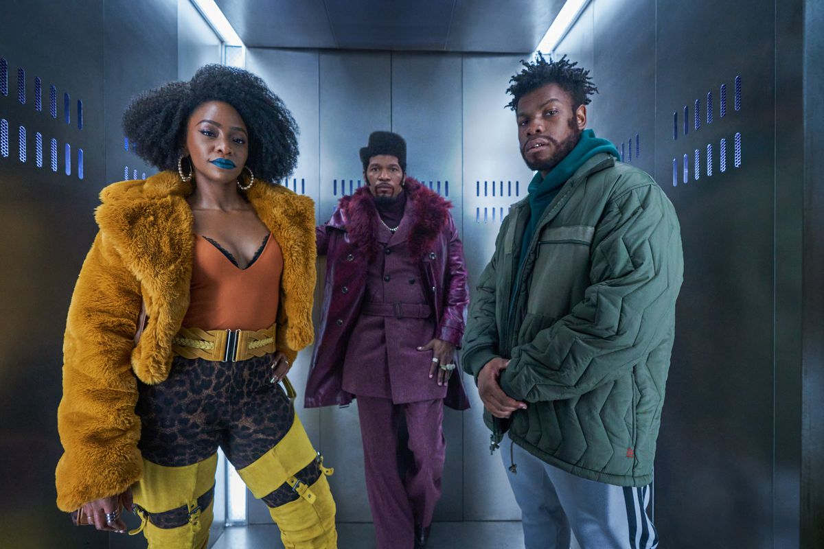 (De gauche à droite) Teyonah Parris dans un manteau de fourrure orange, un body orange avec des peintures à imprimé léopard, Jamie Foxx dans un costume entièrement violet avec un manteau assorti et John Boyega dans un manteau bleu sarcelle gonflé debout dans un ascenseur métallique dans Ils ont cloné Tyrone.