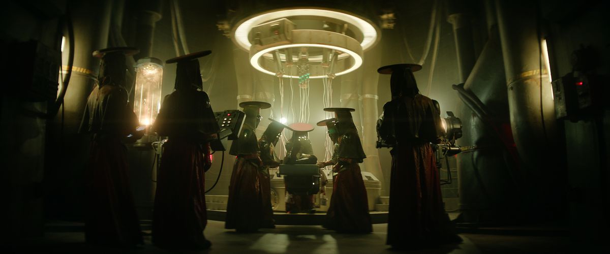 Un groupe de personnages mystérieux vêtus de robes rouges, de chapeaux plats et de voiles entourent un objet sous un dispositif circulaire et annelé suspendu au plafond dans Rebel Moon Part One: A Child of Fire.