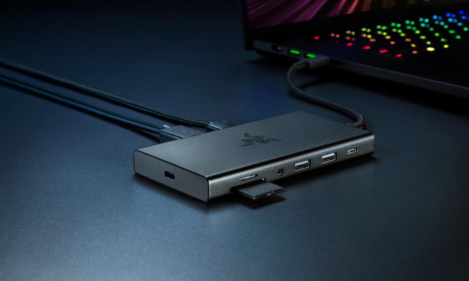 Photo marketing du produit de la station d'accueil Razer USB C.  Le hub se trouve sur un bureau avec un ordinateur portable de jeu derrière lui.  Il dispose de plusieurs ports ouverts et d’une carte SD à mi-chemin.  Il repose sur un bureau bleu foncé avec des ombres spectaculaires.