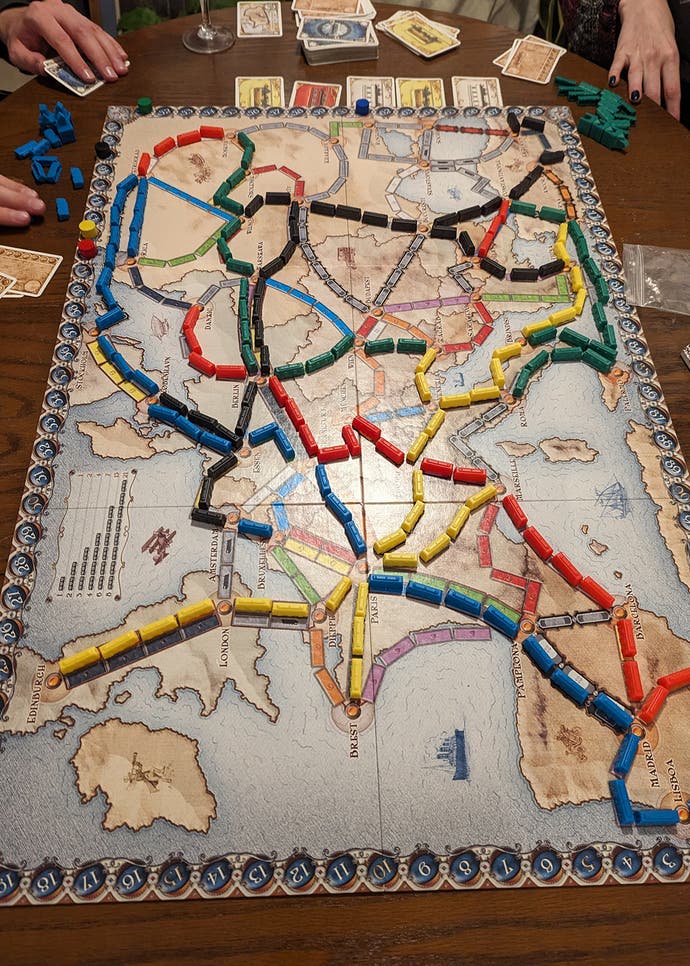 Une photo du jeu de société Ticket to Ride en cours de lecture.  Il s'agit d'une image de haut en bas montrant le plateau du jeu, semblable à une carte, entouré de trains en plastique de couleur primaire qui serpentent tout autour.