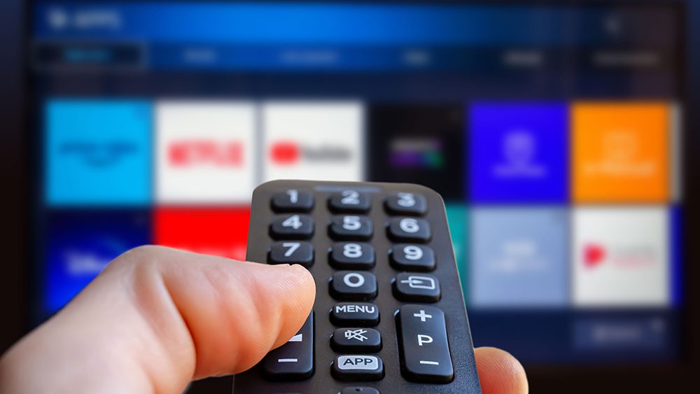 A man is holding a remote control of a smart TV in his hand. In the background you can see the television screen with streaming entertainment apps for video on demand