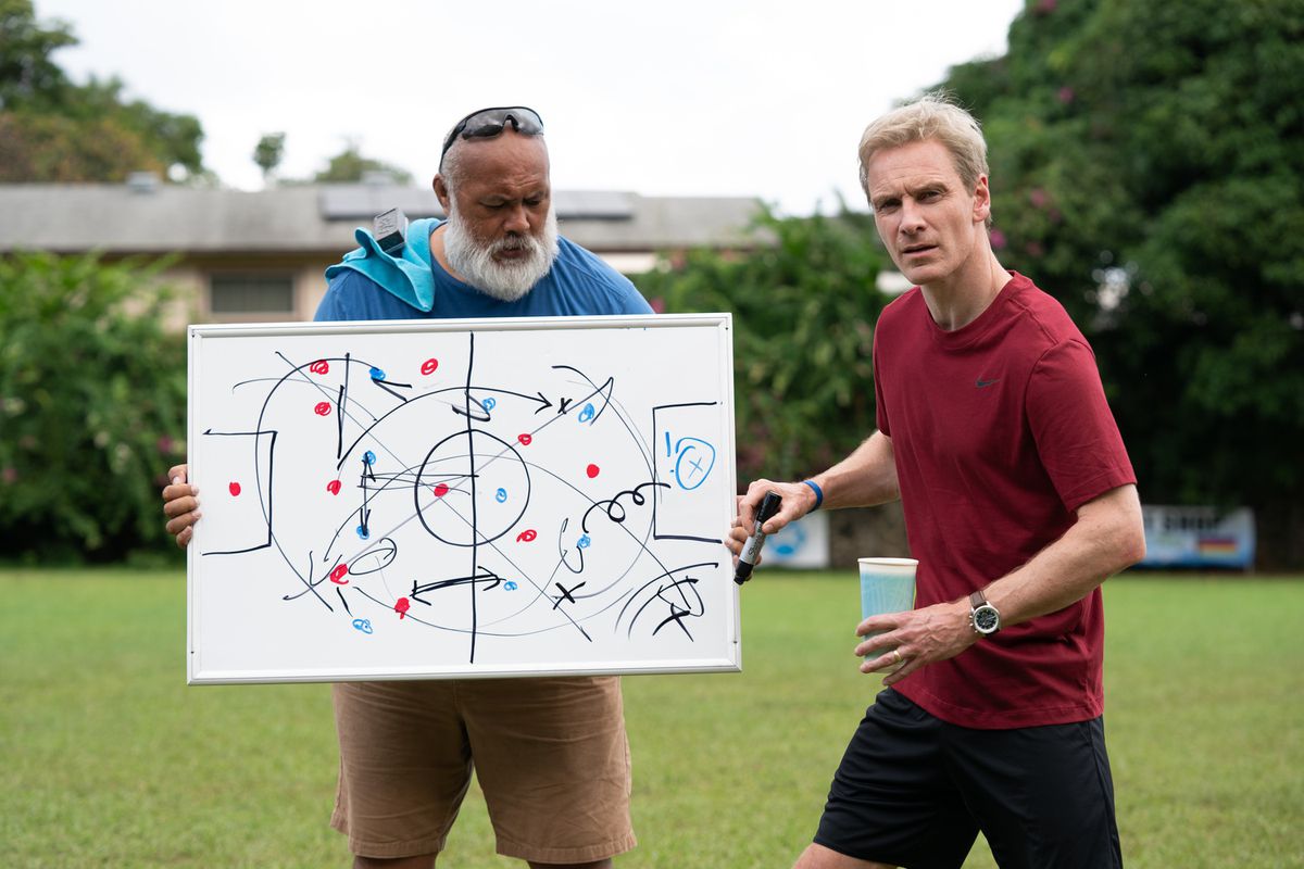 Ace (David Fane) tenant un tableau blanc pendant que l'entraîneur Thomas Rongen (Michael Fassbender) donne une conférence à son équipe hors écran dans Next Goal Wins.