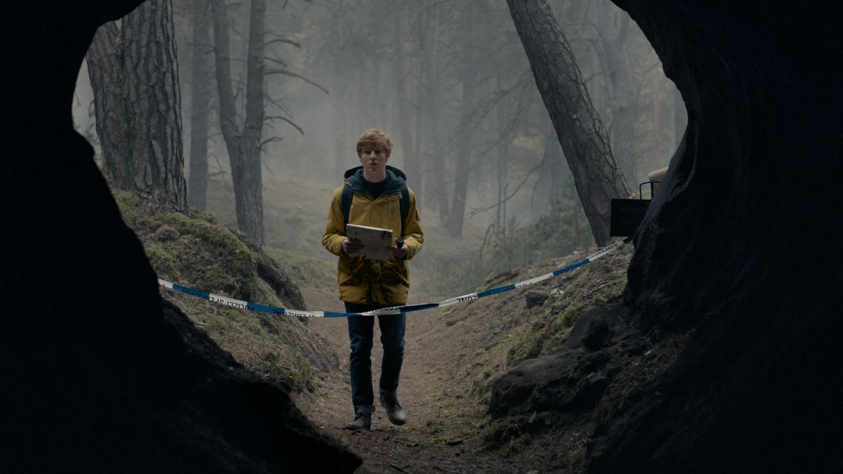 Un jeune homme vêtu d'un imperméable jaune se tient devant l'entrée cloisonnée d'une grande grotte dans une forêt de Dark.