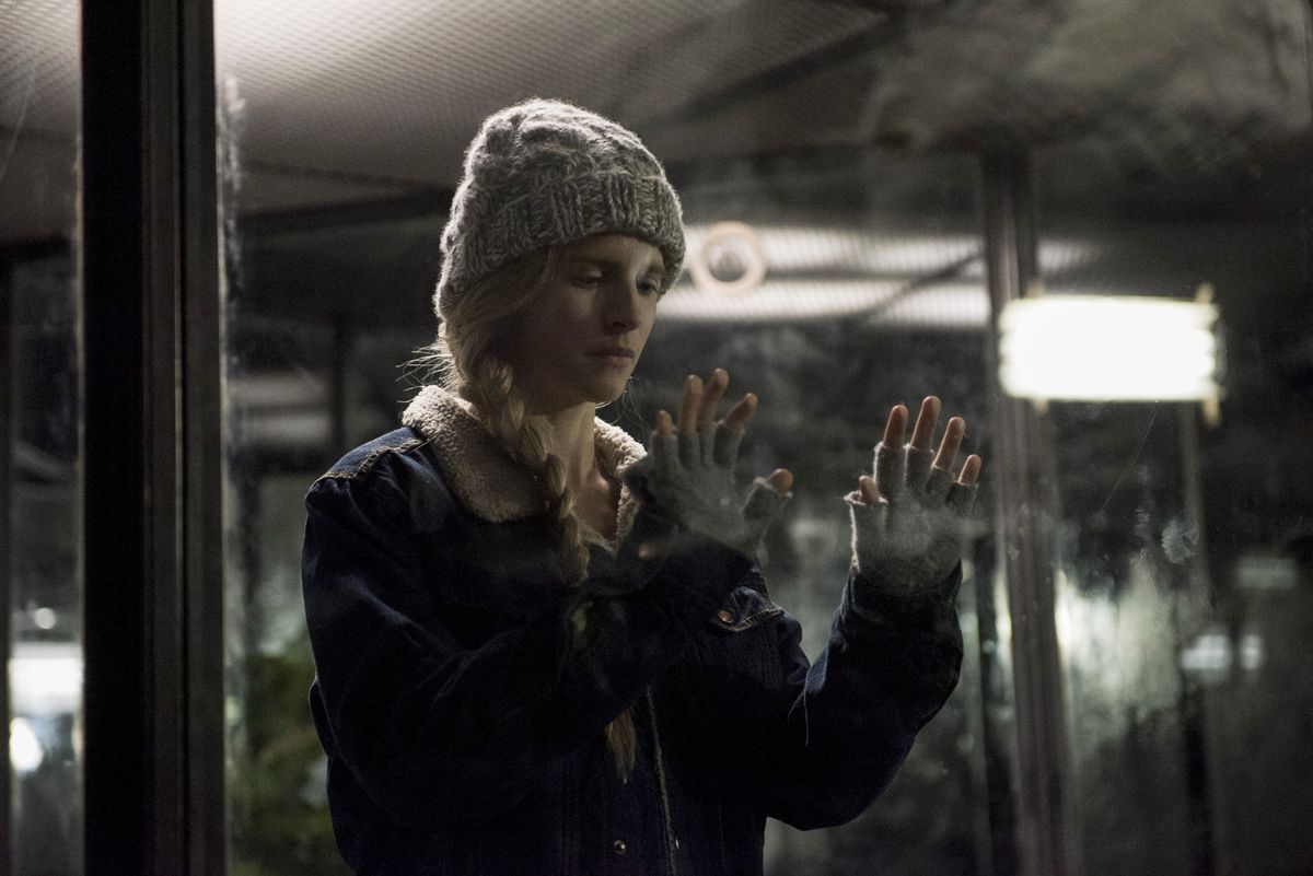 Prairie Johnson dans un manteau, une casquette en tricot et des gants sans doigts pressant ses mains contre une vitre tachée dans The OA.