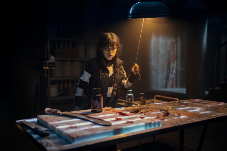 Wednesday Addams (Jenna Ortega) stands in front of a creepy, bloody work table in episode 106 of Wednesday