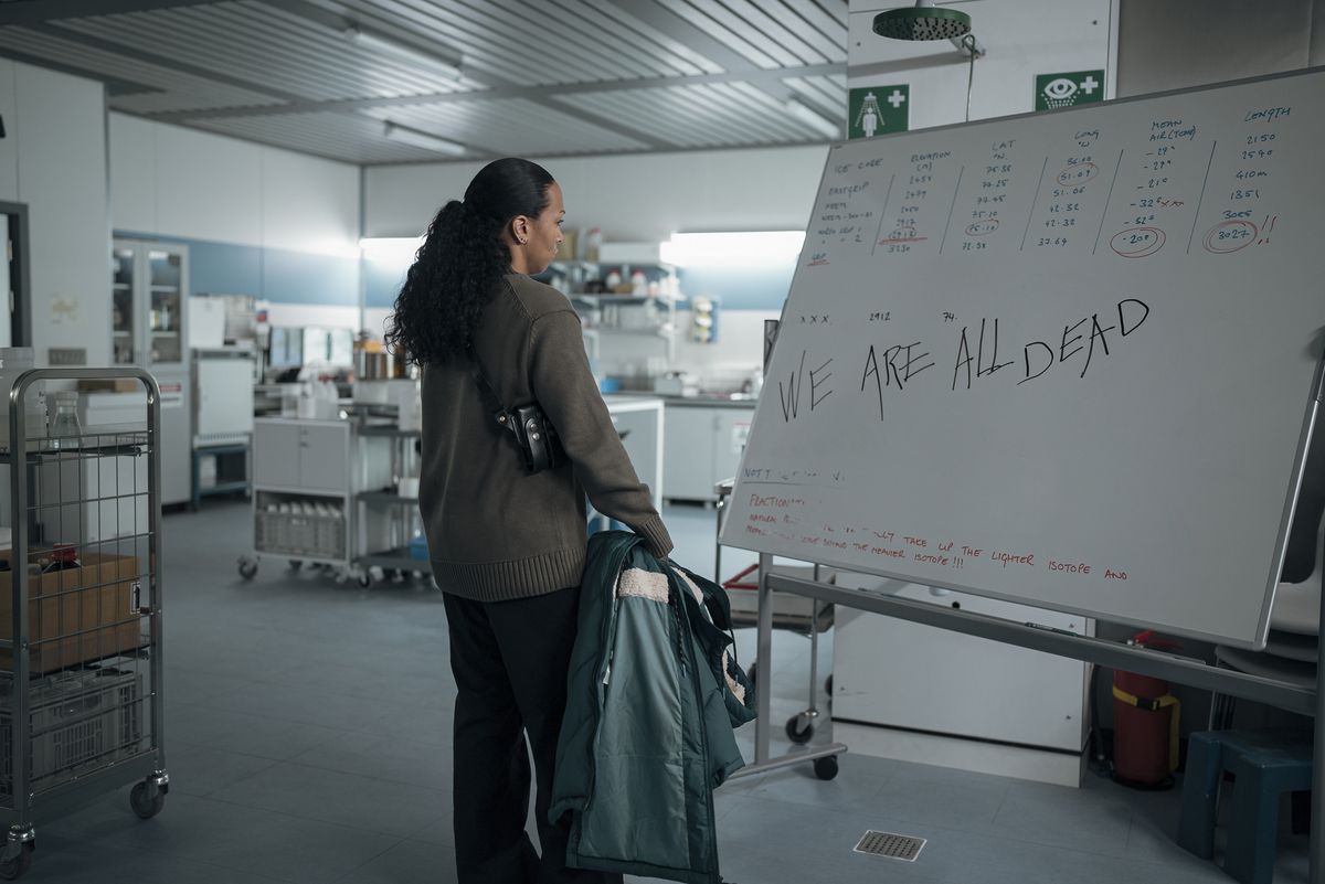 Kali Reis dans le rôle d'Evangeline Navarro se tenant devant un tableau partiellement effacé avec les mots « Nous sommes tous morts » dans True Detective : Night Country.