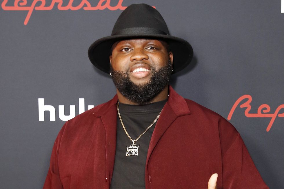 wavyy jonez, un homme sourit à la caméra, il a une barbe noire, il porte un haut et un pantalon noirs avec une chemise rouge et un chapeau noir