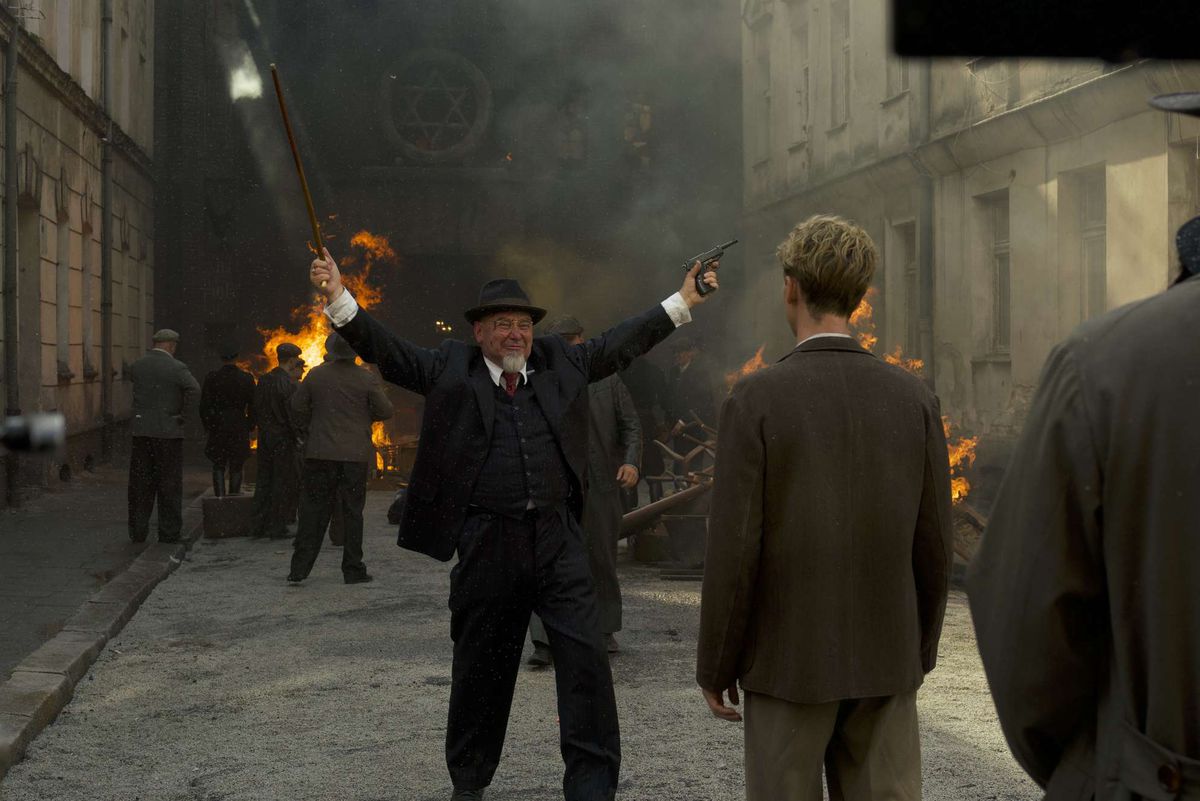 Un homme avec un chapeau et une barbe blanche pointue, sans moustache, lève les bras en triomphe devant une synagogue en feu.  Il tient une arme à feu