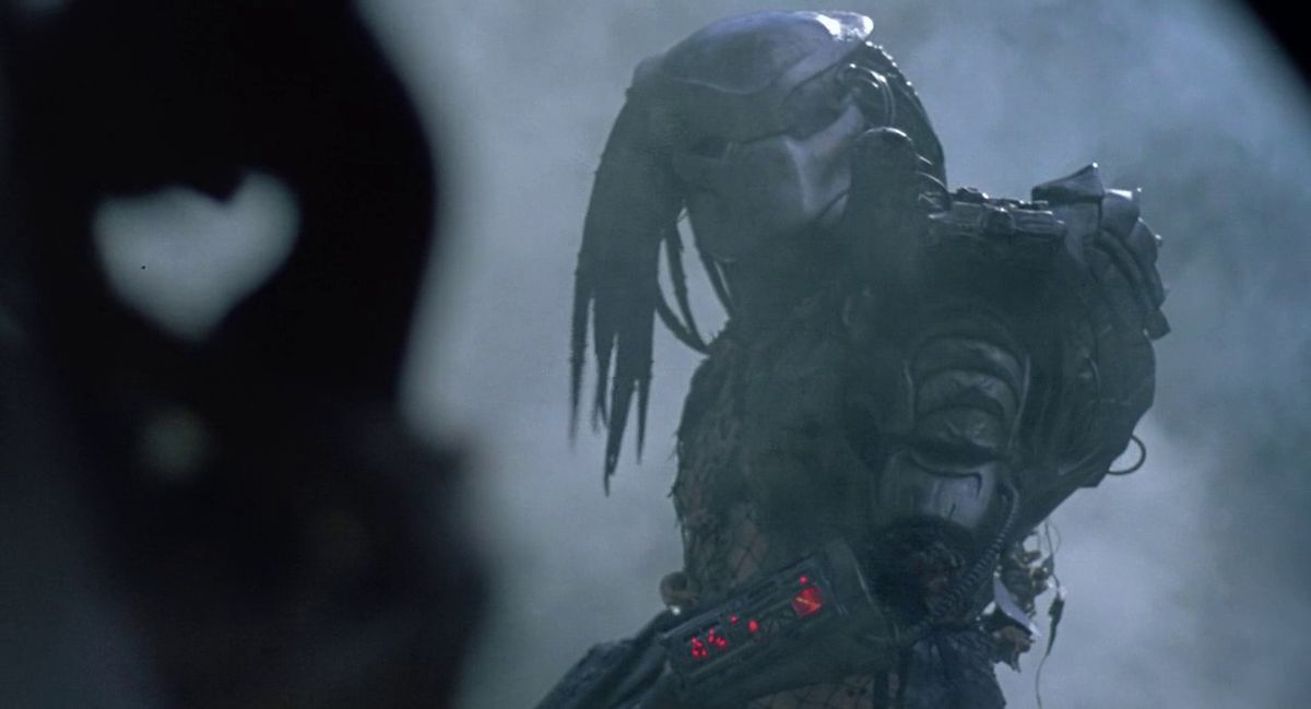 Un guerrier extraterrestre en armure portant un masque avec des dreadlocks se tient dans une jungle entourée de fumée dans Predator.