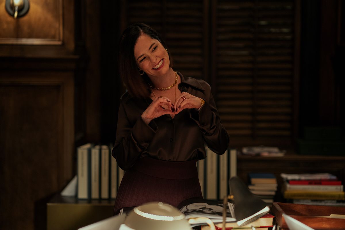 Parker Posey sourit et forme un cœur d'amour avec ses mains dans un bureau sombre de Mr. &  Madame Smith