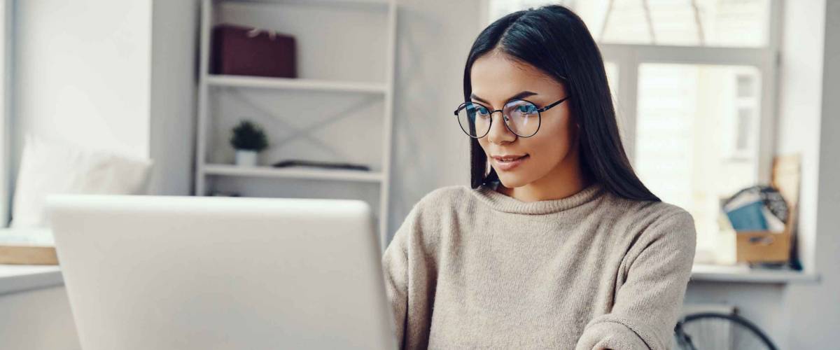 Belle jeune femme vêtue de vêtements décontractés utilisant un ordinateur portable et souriant tout en travaillant à l'intérieur