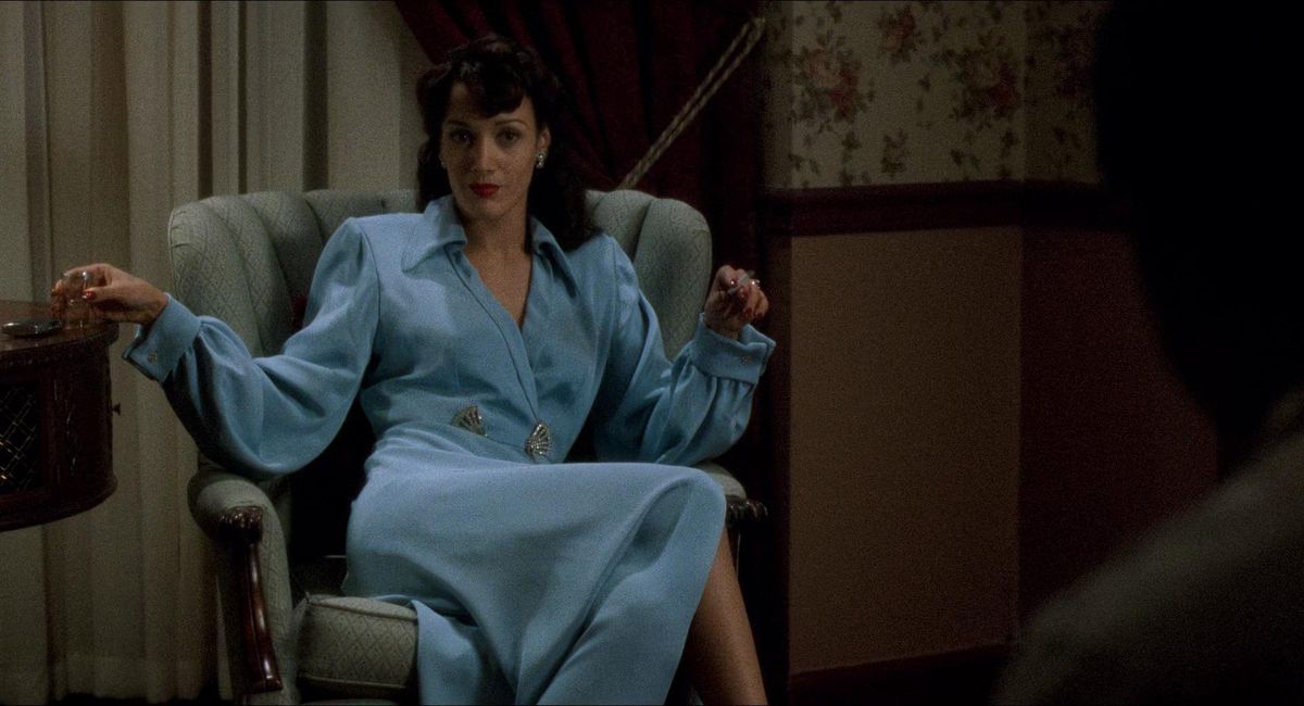 Jennifer Beals porte une robe bleu clair alors qu'elle est assise sur une chaise dans Le Diable en robe bleue (c'est le diable en robe bleue, dit votre rendez-vous)