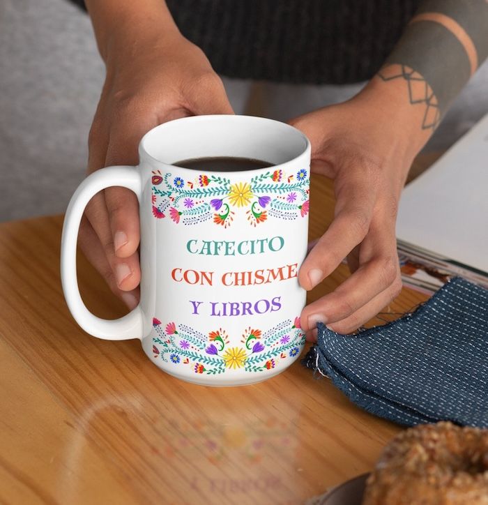 une tasse avec un motif floral peint en haut et en bas.  Entre les fleurs se trouvent les mots "cafécito avec chisme et livres"